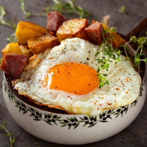 Egg with fries and fried salami in a rustic pan with some herbs